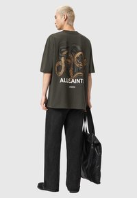 Oliwkowa koszulka z krótkim rękawem z grafiką węża i napisem "ALLSAINTS LONDON" na plecach, połączona z czarnymi szerokimi spodniami i czarną torbą.