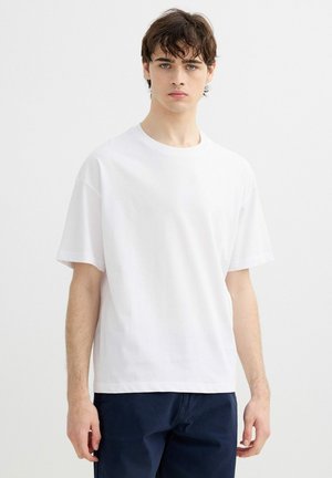 Joven con cabello corto y oscuro, que lleva una camiseta blanca lisa de manga corta y pantalones oscuros, de pie frente a un fondo claro y neutro.