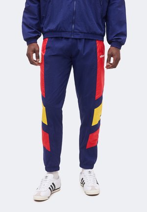 Mężczyzna w granatowych spodniach dresowych z czerwonymi i żółtymi panelami geometrycznymi oraz białych butach sportowych Adidas z czarnymi paskami.