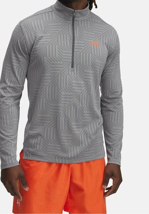 Mann trägt ein graues, gemustertes Langarm-Quarter-Zip-Shirt und leuchtend orangefarbene Sportshorts, steht mit entspannten Händen an den Seiten.