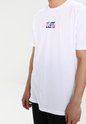 T-shirt blanc à manches courtes avec un petit logo Diesel coloré sur la poitrine, porté par une personne debout devant un fond blanc uni.