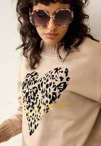 Béžová mikina s velkým černým leopardím potiskem ve tvaru srdce a zlatým písmem. Límec je žebrovaný a látka působí měkce.