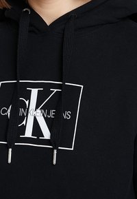 Sudadera negra con capucha de algodón, con un logo gráfico blanco que incluye el texto "CK" y "CALVIN KLEIN JEANS" debajo. Incluye cordones ajustables.