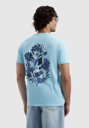 Man met krullend haar die een lichtblauw T-shirt draagt met een donkerblauw bloemmotief en witte letters op de achterkant, gecombineerd met licht denim korte broek.