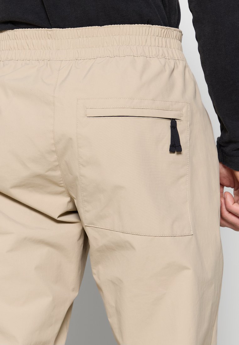 Pantalon beige avec ceinture élastique et poche arrière dotée d'une fermeture éclair noire, porté avec un haut noir à manches longues.