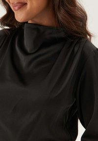 Top in tessuto nero e liscio con scollatura a drappeggio e maniche lunghe. Presenta una texture sottile e morbida e un design minimalista.