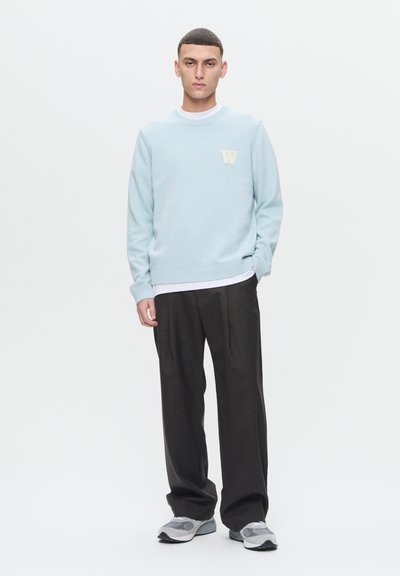 Světle modrý pletený svetr s žebrovanými manžetami, se smetanovou emblematickou kanceláří 'W' na hrudi. V kombinaci s volnými tmavými kalhotami a teniskami.