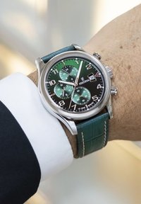 Reloj con correa de cuero verde, caja de acero inoxidable, esfera negra, acentos plateados y cuatro sub-esferas verdes; cuenta con un indicador de fecha.