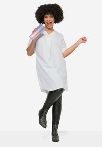 Witte oversized shirt met korte mouwen, gecombineerd met zwarte legging en met studs versierde enkelboots, terwijl hij/zij een paarse gradient mok vasthoudt.
