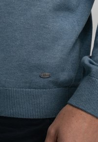  Lichtblauw gebreide trui met een geribbelde rand en manchetten, met een klein, ovaal metalen logo op de onderste linkerkant.