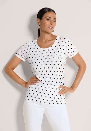 Frau mit dunklen Haaren, die goldene Creolen trägt und ein weißes Kurzarmhemd mit schwarzen Punkten, posiert mit den Händen in der Hüfte.