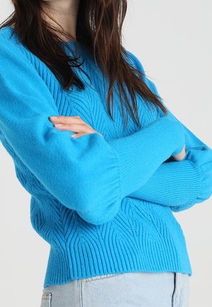 Femme aux longs cheveux bruns portant un pull bleu vif à torsades avec des manches bouffantes, bras croisés, et un jean bleu clair.