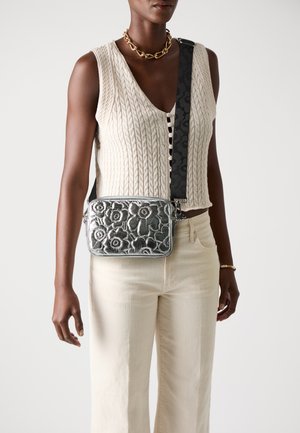 Geantă crossbody metalică argintie cu un design embossat floral, asociată cu o curea neagră cu model, purtată peste un top din tricot cu cablu și pantaloni bej.