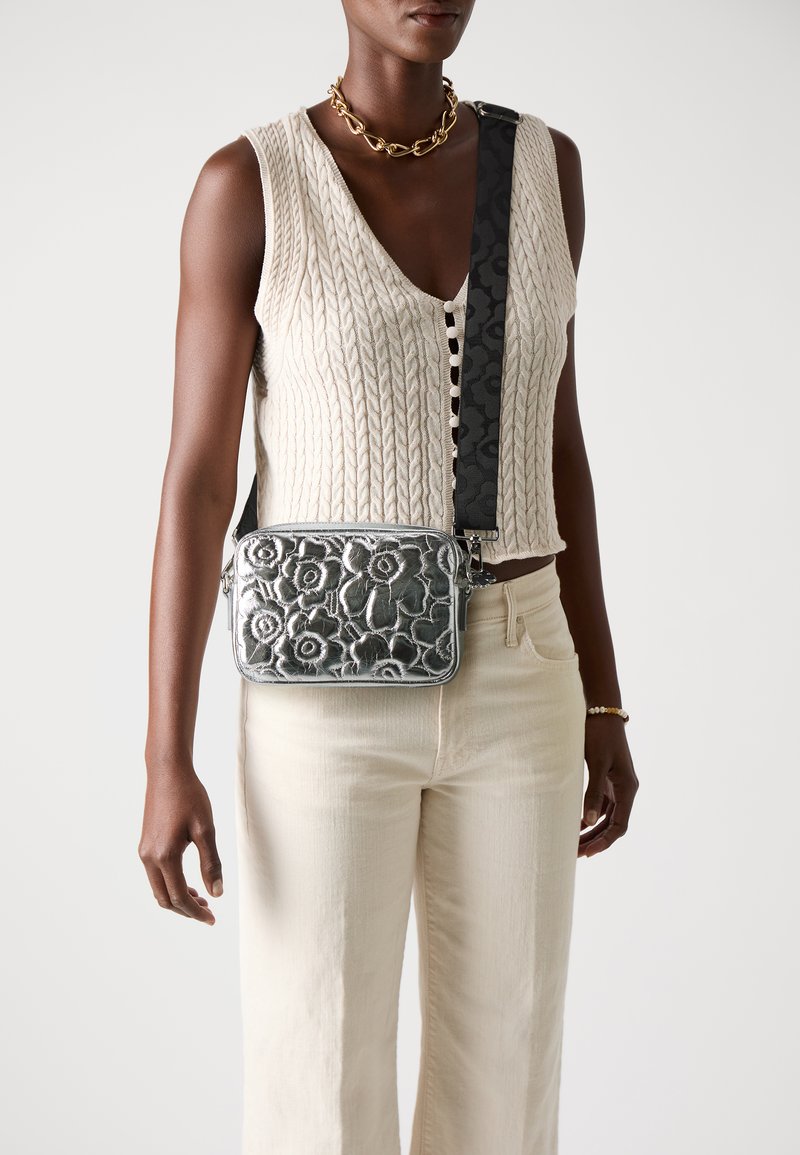 Bolso cruzado metálico plateado con diseño de flores en relieve, acompañado de una correa negra con estampado, llevado sobre un top de punto trenzado y pantalones beige.