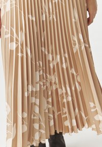 Jupe midi plissée en beige avec un motif de feuilles blanches. Fabriquée en tissu doux, elle présente des plis verticaux et une texture lisse.