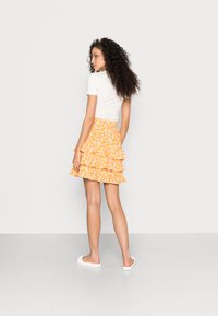 Top branco de mangas curtas acompanhado de uma saia amarela floral em camadas e com babados. A modelo usa sapatos brancos sem atacadores. O fundo é cinza liso.