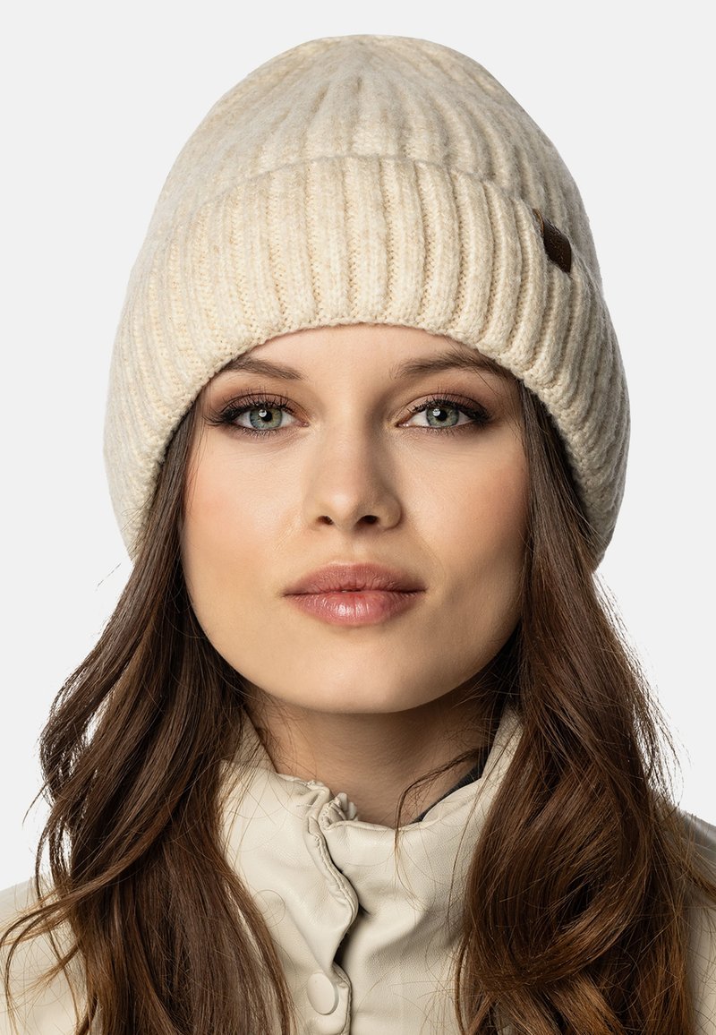 Crème-kleurige gebreide beanie met een geribde textuur, voorzien van een klein bruin leren accent. Zacht materiaal en een afgeronde kroonontwerp.