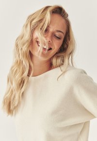 Cremefarvet strikket sweater med en afslappet pasform, bred halsudskæring og blød tekstur, med lange, bølgede blonde hår, der falder over skulderen.