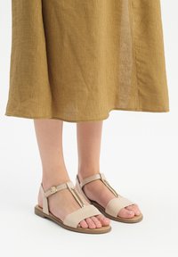 Vaaleanbeige tasapohjaiset sandaalit T-mallin vyöllä, joissa on ketjuakseli, säädettävä nilkkavyö ja teksturoitu päällinen.