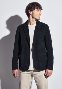 Černý kordový blejzr s klopami, dvěma předními kapsami a knoflíkovým zapínáním. Nositý přes světlejší tričko a spárovaný s krémovými kalhotami.