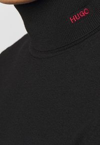 Maglione nero a collo alto in un tessuto a maglia liscia, con un colletto a coste alto e un piccolo logo rosso "HUGO" sul collo.
