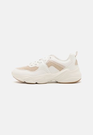 Zapatilla gruesa blanca y beige con paneles de malla y cuero, suela gruesa, cierre de cordones y lengüeta en el talón, vista de lado.