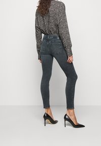 Jeans de mezclilla oscura de cintura alta con un corte ajustado, combinados con zapatos de tacón stiletto de charol negro. Una blusa de patrón texturizado completa el conjunto.