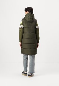 Polstret lang vest i mørkegrønn med hette, med kviltet design og avslappet passform, kombinert med en mønstret strikkegenser og jeans.