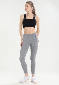 Sujetador deportivo negro con tirantes con logo, combinado con leggins de cintura alta en gris claro que presentan costuras laterales. La modelo lleva deportivas blancas.