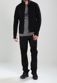 Man som bär en svart softshelljacka, en grå t-shirt med grafik, svarta utemuggar och svarta vandringsskor, står mot en neutral bakgrund.