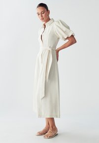 Crème-kleurige linnen jurk met een aansluitende lijfje, pofmouwen, een ceintuur in de taille en een midi-lengte. Gecombineerd met minimalistische platte sandalen.