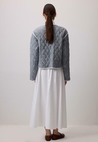 Prešívaná sivá bunda s kvetinovou výšivkou a bielym lemom, nosená cez dlhú bielu sukňu. Hnedé slip-on topánky dopĺňajú outfit.