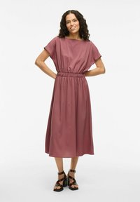 Mauve obleka s kratkimi rokavi in elastičnim pasom, ki se razteza v elegantno A-linijo, kombinirana s črnimi sandali na peto z naramnicami.
