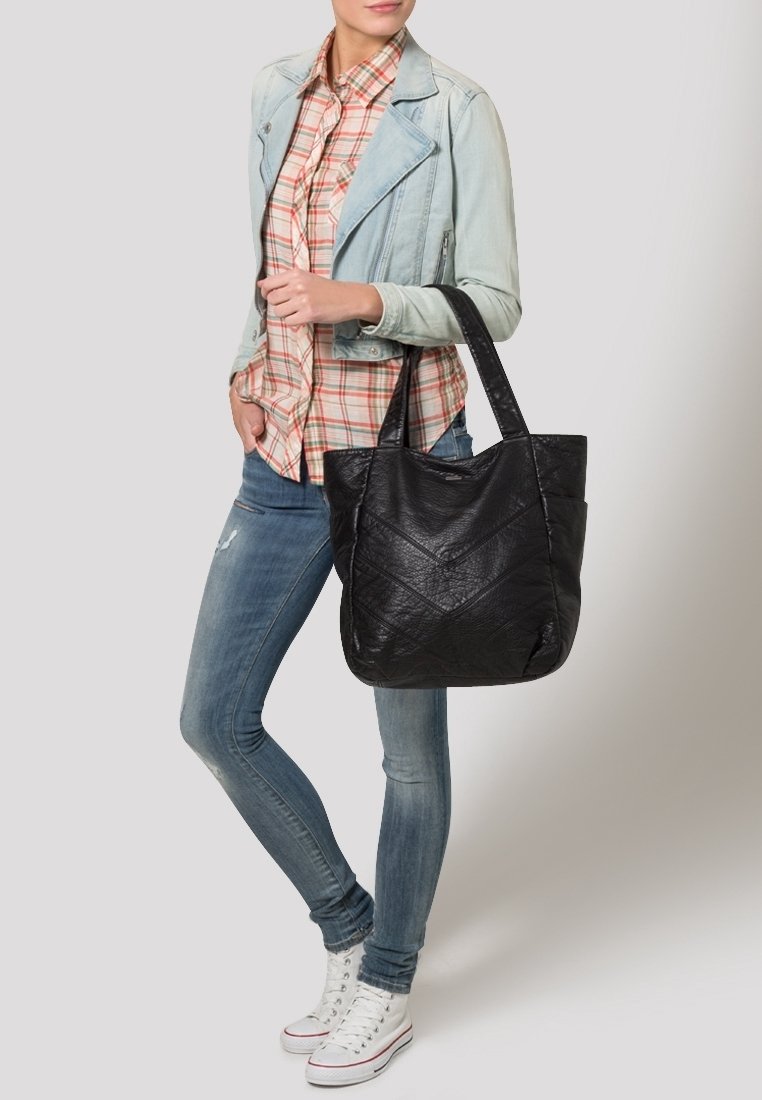 Femme portant une chemise à carreaux, une veste en jean claire, un jean slim et des baskets blanches, tenant un grand sac cabas en cuir noir sur son épaule.