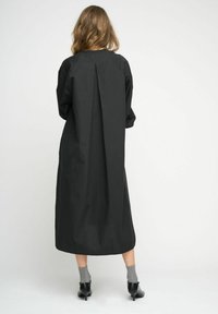 Rochie neagră midi, cu croială relaxată, detalii cu pliuri la spate și mâneci lungi. Purțată cu șosete gri și pantofi cu toc. Textură netedă.
