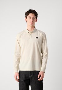 Langermet polo-skjorte i lys beige, med knappet krage, en liten logo-patch på brystet og ribbestrikkede mansjetter. Kombinert med mørke jeans.