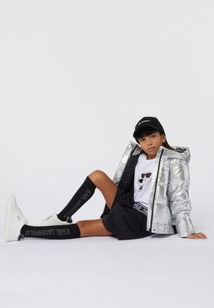 Zilveren pufferjack met een capuchon, wit graphic t-shirt, zwarte shorts, kniehoge zwarte sokken met witte tekst en witte sneakers.