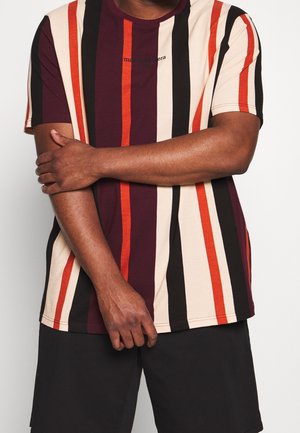 Mężczyzna ubrany w koszulę w pionowe paski w kolorach bordowym, kremowym, czarnym i pomarańczowym, z założonymi rękami, w czarnych szortach na białym tle.