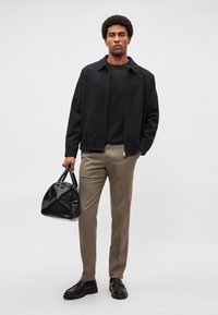 Musta vetoketjullinen takki mustan paidan päällä, beiget housut ja mustat kengät. Pitää kädessään kuviollista mustaa käsilaukkua. Yksinkertainen, jäsennelty muotoilu.