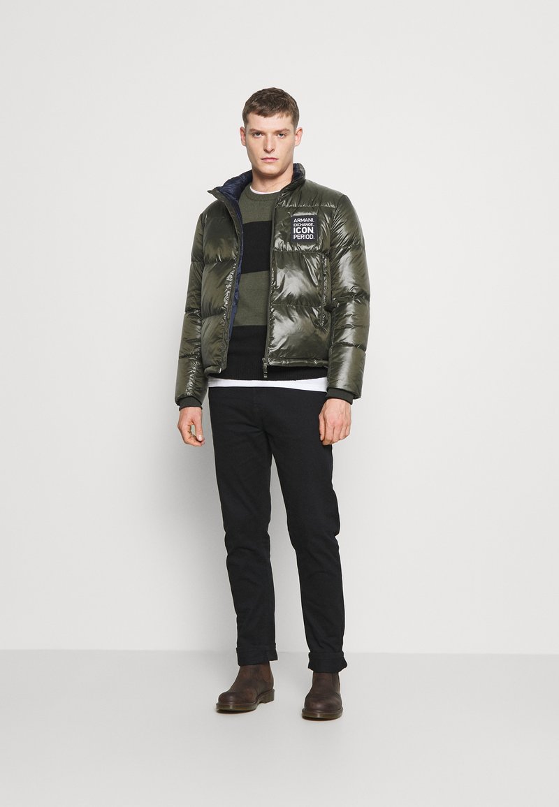 Giubbotto imbottito verde oliva con una finitura lucida, chiusura con zip e dettaglio del logo sul petto. Abbinato a un maglione scuro e pantaloni neri.