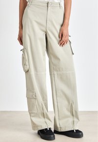 Cargo pants in light beige cotton, featuring large side pockets, zippered accents at the ankles, and a relaxed fit. Black shoes with laces.