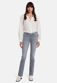 Blouse blanche à manches longues et boutons avec des détails en dentelle, associée à un jean gris clair ajusté et à des mocassins crème, se tenant devant un fond blanc.