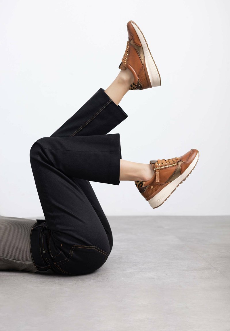 Zapatillas de cuero marrón con acentos beige, con cordones en la parte frontal y una suela media texturizada. Pantalones de mezclilla negra con costuras en contraste.