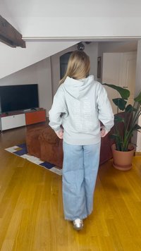 Ljusgrå hoodie med text på baksidan, kombinerad med vida blå jeans. Rummet har trägolv, krukväxt och en TV.