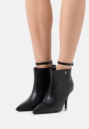 Botas de cuero negras hasta el tobillo con punta afilada, tacón alto y una correa delgada en el tobillo con detalles en plata. Textura suave en toda su superficie.