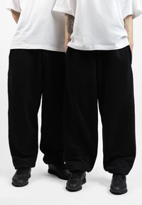 Black wide-leg sweatpants with a textured finish, worn with black sneakers. Two individuals display pants and loose-fitting white t-shirts.