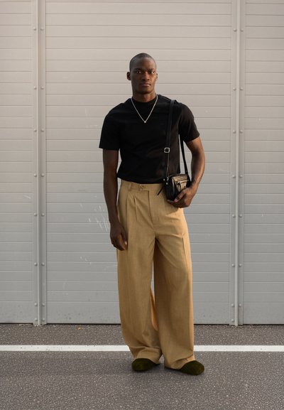 Μαύρο πουκάμισο με κοντά μανίκια, μπεζ παντελόνι με ευρεία μπατζάκια, μαύρη τσάντα χιαστί, πράσινα παπούτσια, όρθιος μπροστά σε φόντο γκρι πάνελ.