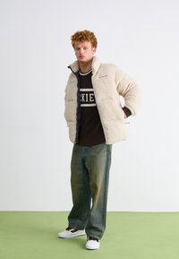 Puffer jacket in cream fleece with zip closures and black lining, worn over a brown sweater with white text, paired with light blue jeans and sneakers.