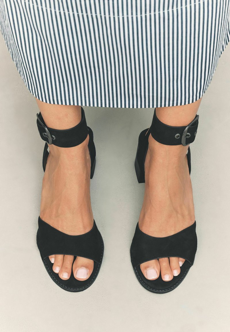Schwarze High-Heels-Sandalen mit Knöchelriemen, kombiniert mit einem gestreiften Rock, der gepflegte Füße mit lackierten Zehennägeln hervorhebt.