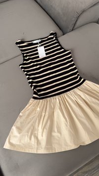 Mouwloze jurk met zwarte en beige horizontale strepen bovenaan en een beige geplooide rok, plat gelegd op een grijze bank.
